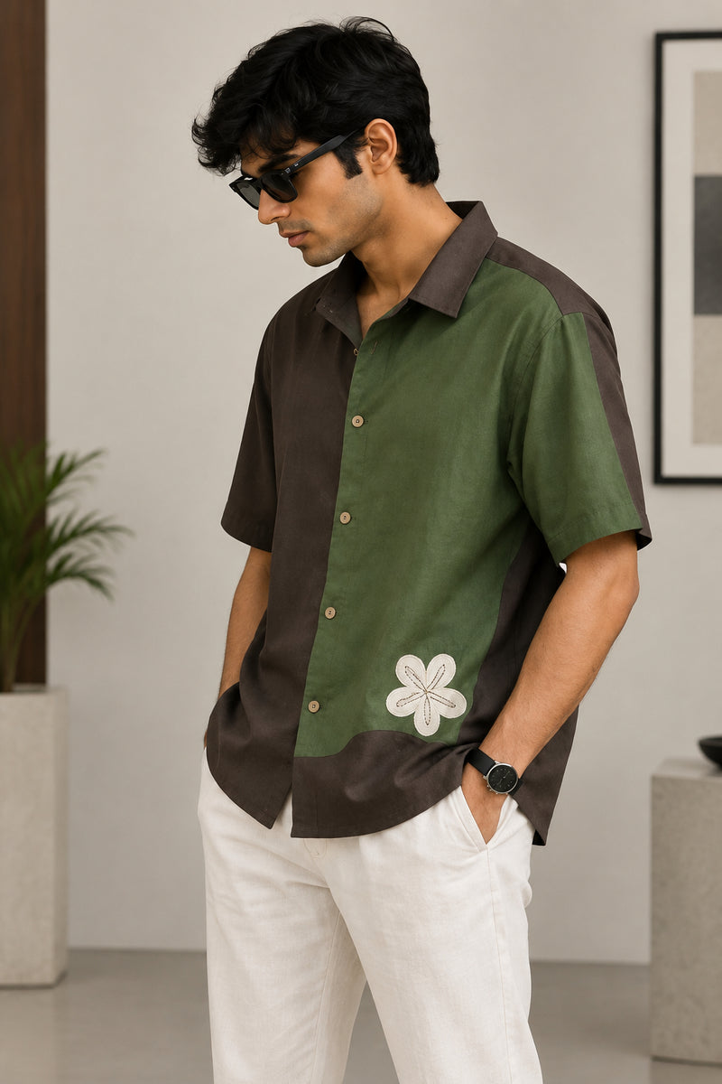 A man wearing a short-sleeve olive green and dark brown button-up shirt with a  embroidered flower on the front.