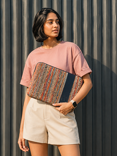 A woman holds a laptop sleeve with a multicolor striped pattern and a navy blue  organic leather.