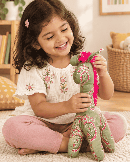 Child sitting on a rug playing with a green paisley fabric horse plush toy with a pink fringe mane
