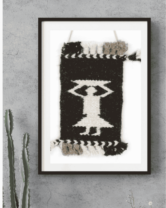 A framed black and white woven wall hanging with a geometric figure design, displayed on a textured gray wall next to a cactus.